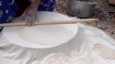 Mavi elbiseli kadın hamuru beyaz bir masa örtüsüne sarar. Elinde tahta bir oklavayla geleneksel düz ekmek hazırlar.