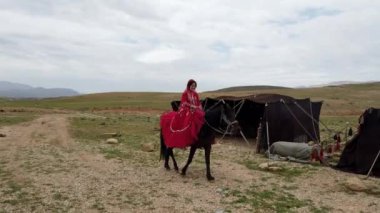 Kırmızı elbiseli bir kadın, dağlık bir bölgede çadırların yanından ata biniyor ve İran 'ın göçebe yaşam tarzını gösteriyor.