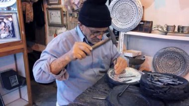 Şapka ve gözlük takan deneyimli bir zanaatkar dikkatlice bir çekiç ve keski kullanarak bir tasarımı metal bir plakaya kazıyor.