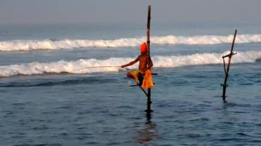 Turuncu türbanlı Sri Lankalı balıkçı bir çubuk üzerinde denge kurarken ipini okyanusa fırlatıyor.