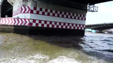 Bangkok 'ta bir nehir boyunca hızla giden yolcularla dolu kanal teknesi, dinamik bir hareketle köprünün altından geçiyor.