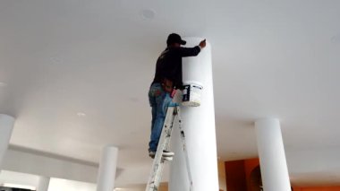 İnşaat işçisi, büyük beyaz bir sütunu boyarken bir merdivenin üzerinde duruyor, detaylara dikkat ve beceri gösteriyor.