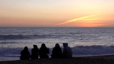 Beş arkadaş, kumsalda oturup okyanusun üzerinde günbatımını izleyerek huzurlu bir anın tadını çıkarıyor.
