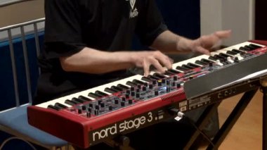 Siyah tişörtlü bir müzisyen sahnede kırmızı bir klavye sentezleyicisi çalıyor, müzik için yetenek ve tutku sergiliyor.