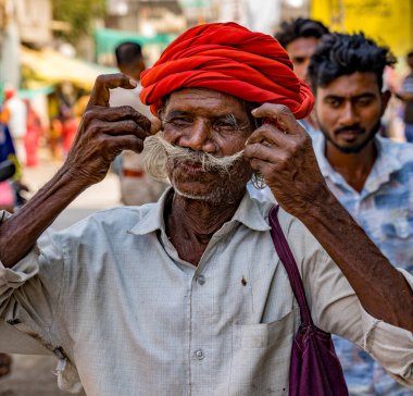 Kıdemli, kırmızı sarıklı Hintli adam, kalabalık bir sokak sahnesinde bıyığını düzeltiyor.