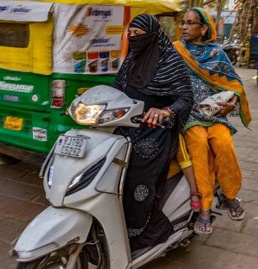 İki Hintli kadın, biri geleneksel giyimli, canlı bir sokak ortamında scooter kullanıyorlar.