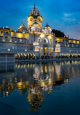 Aydınlatılmış altın tapınağın yansıtıcı alacakaranlık görüntüsü ve kutsal havuz boyunca dindarlar.