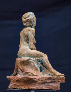 Küçük terracotta heykelciği, bir kayanın üzerinde oturan stilize edilmiş bir kadın figürünü temsil ediyor, arkadan görülüyor, koyu bir arka planda.