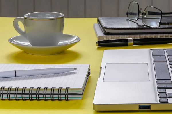 Laptop, notebooks with pens and a cup of coffee on a desktop with a yellow surface