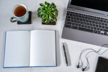 Kalem ve dizüstü bilgisayarı olan boş bir defter, akıllı telefon ve beyaz masaüstünde bir fincan çay