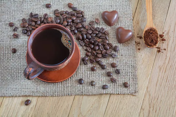 Coffee with foam in a brown clay cup, coffee beans and two heart-shaped candies on a wooden table