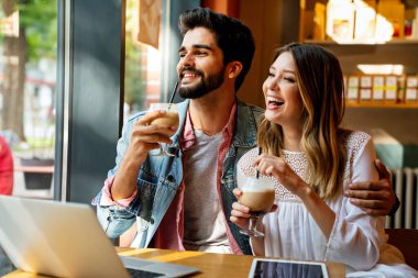 Kafede oturmuş gülümseyen, dizüstü bilgisayara bakan, keyifle vakit geçiren, kahve içen mutlu genç bir çift..