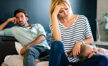 Tired young mother and father sitting on couch feels annoyed exhausted stressful with little girl child.