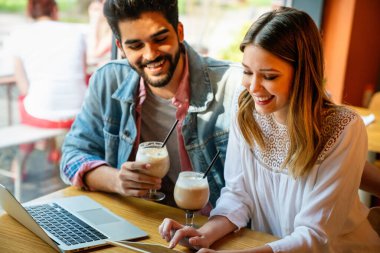Kafede oturmuş gülümseyen, dizüstü bilgisayara bakan, keyifle vakit geçiren, kahve içen mutlu genç bir çift..