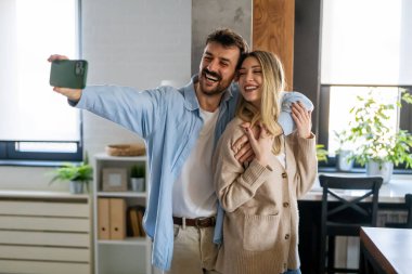Genç, çekici, gülümseyen, etnik çeşitlilikte bir çift modern evde cep telefonuyla selfie çekiyor.