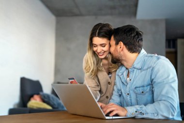 Mutlu genç çift evde online alışveriş yaparken dizüstü bilgisayar kullanıyor ve gülümsüyor.