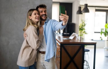 Genç, çekici, gülümseyen, etnik çeşitlilikte bir çift modern evde cep telefonuyla selfie çekiyor.