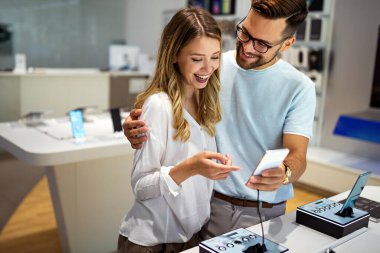 Genç ve tatmin olmuş bir çift, teknoloji mağazasının masasındaki cep telefonunun yeni modelini test ediyor..