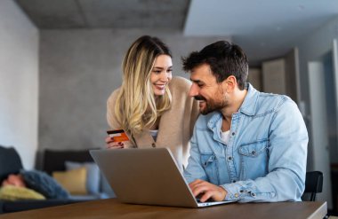 Mutlu genç çift evde online alışveriş yaparken dizüstü bilgisayar kullanıyor ve gülümsüyor.