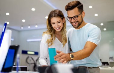 Genç ve tatmin olmuş bir çift, teknoloji mağazasının masasındaki cep telefonunun yeni modelini test ediyor..