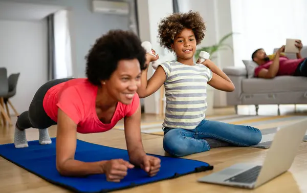Beautiful black mother and teen daughter having online yoga class together, making exercises on fitness mat in front of laptop