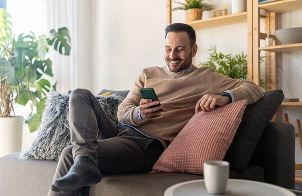 Привлекательный молодой счастливый мужчина отдыхает дома на диване, пользуется мобильным телефоном, пьет кофе