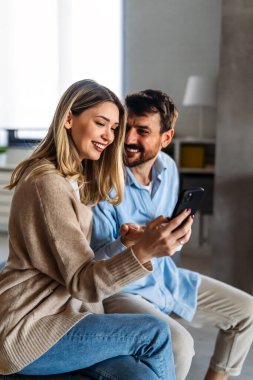 Akıllı telefona bakarken kucaklaşan gülümseyen genç çift. Sosyal medyayı akıllı telefondan paylaşan çok ırklı çift.