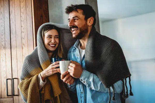 Happy young couple with hot drinks cuddling under warm blanket at home. Weekend people relax love concept.