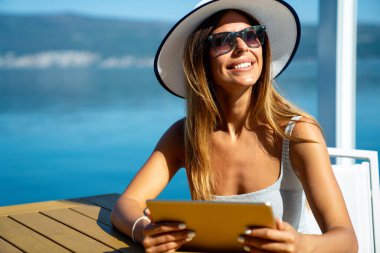 Rahat ve neşeli. İş ve tatil. Deniz kenarındaki terasta tablet bilgisayar kullanan mutlu genç bir kadının açık hava portresi..