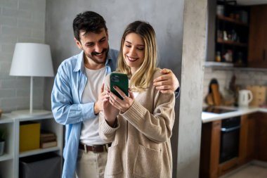 Genç mutlu çift evde sosyal medya haberlerini paylaşmak için telefonu kullanıyor, gülümsüyor, internetten alışveriş yapıyor. Mobil teknoloji yaşam tarzı kavramı