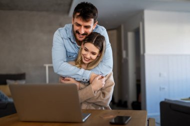 Mutlu genç çift evde online alışveriş yaparken dizüstü bilgisayar kullanıyor ve gülümsüyor.