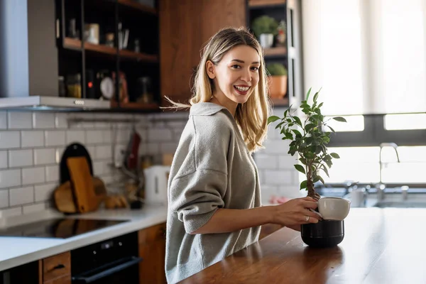 Smiling young beautiful woman holding can, watering green plants in pot. Happy attractive housewife enjoying taking care of domestic flowers, gardening and housekeeping hobby concept.