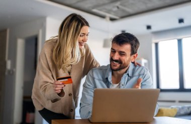 Mutlu genç çift evde online alışveriş yaparken dizüstü bilgisayar kullanıyor ve gülümsüyor.