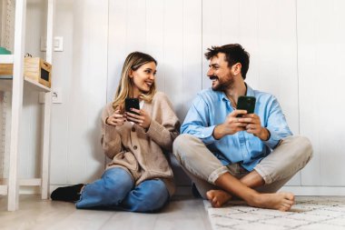 Evde akıllı telefondan birlikte gülümseyen bir çift. İki telefon kullanan genç mutlu insanlar evde sosyal medya haberlerini paylaşıyorlar