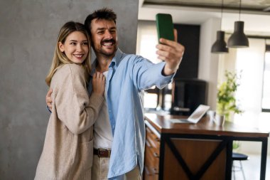 Genç, çekici, gülümseyen, etnik çeşitlilikte bir çift modern evde cep telefonuyla selfie çekiyor.