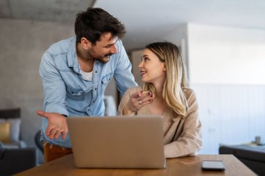 Mutlu genç çift evde online alışveriş yaparken dizüstü bilgisayar kullanıyor ve gülümsüyor.