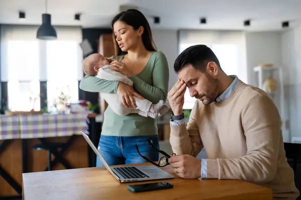 Father businessman try to work on laptop from home with wife and baby on background. Work from home concept. Freelancer