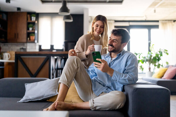 Young happy couple using phone to share social media news at home, smiling talking doing shopping online. Mobile tech lifestyle concept