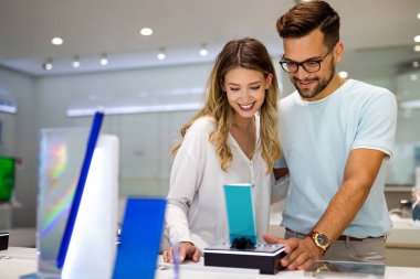 Genç ve tatmin olmuş bir çift, teknoloji mağazasının masasındaki cep telefonunun yeni modelini test ediyor..