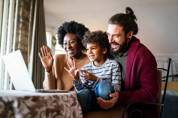 Happy modern multiethnic diverse parents and kid waving hands while using laptop for video chat. People technology online communication concept