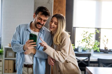 Akıllı telefona bakarken kucaklaşan gülümseyen genç çift. Sosyal medyayı akıllı telefondan paylaşan çok ırklı çift.