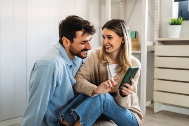 Genç mutlu çift evde sosyal medya haberlerini paylaşmak için telefonu kullanıyor, gülümsüyor, internetten alışveriş yapıyor. Mobil teknoloji yaşam tarzı kavramı