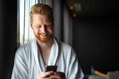 Mutlu gülümseyen adam cep telefonuna bakarken rahatlıyor. Evdeki e-postaları kontrol etmek için akıllı telefon kullanan bir adam. Akıllı telefondan haber okuyan adam.