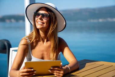 Rahat ve neşeli. İş ve tatil. Deniz kenarındaki terasta tablet bilgisayar kullanan mutlu genç bir kadının açık hava portresi..
