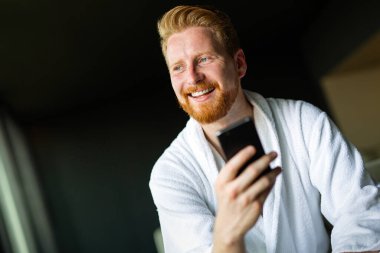 Mutlu gülümseyen adam cep telefonuna bakarken rahatlıyor. Evdeki e-postaları kontrol etmek için akıllı telefon kullanan bir adam. Akıllı telefondan haber okuyan adam.