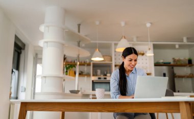 Genç ve güzel bir kadın sosyal medyayı kontrol etmek, çalışmak veya e-postaları okumak için bilgisayar kullanıyor. Dijital cihazlarla evden çalışan yaratıcı iş adamı.