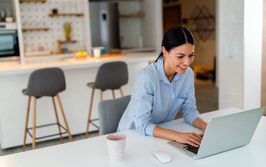 Genç ve güzel bir kadın sosyal medyayı kontrol etmek, çalışmak veya e-postaları okumak için bilgisayar kullanıyor. Dijital cihazlarla evden çalışan yaratıcı iş adamı.