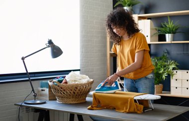 Genç, mutlu, gülümseyen Afro-Amerikan kadın evdeki demir tahtada giysilerini ütülüyor.