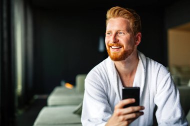 Mutlu gülümseyen adam cep telefonuna bakarken rahatlıyor. Evdeki e-postaları kontrol etmek için akıllı telefon kullanan bir adam. Akıllı telefondan haber okuyan adam.