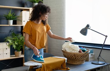 Genç, mutlu, gülümseyen Afro-Amerikan kadın evdeki demir tahtada giysilerini ütülüyor.